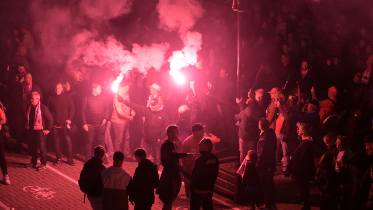 Mensen in een menigte houden fakkels vast die roze rook afgeven, terwijl anderen toekijken.
