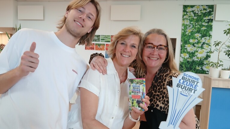Drie mensen poseren vrolijk voor de camera, een van hen houdt een object met tekst "Schone Buurt Leuke Buurt 2026" vast.