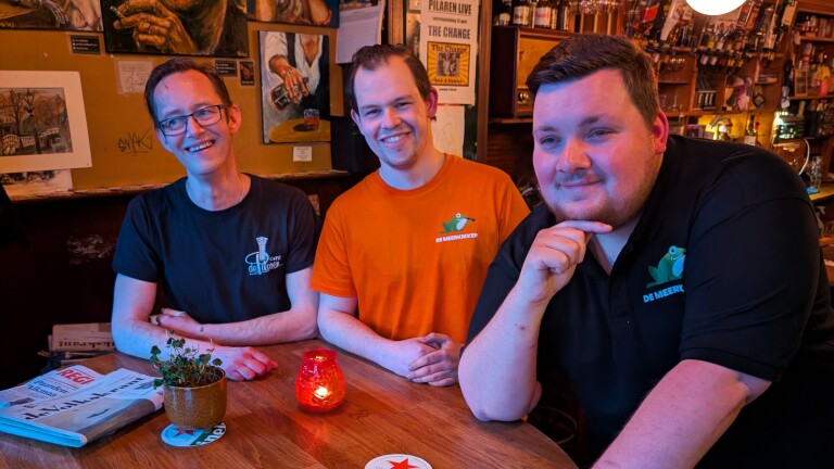 Drie mannen zitten glimlachend aan een tafel in een gezellig café, met schilderijen en drankrekken op de achtergrond.