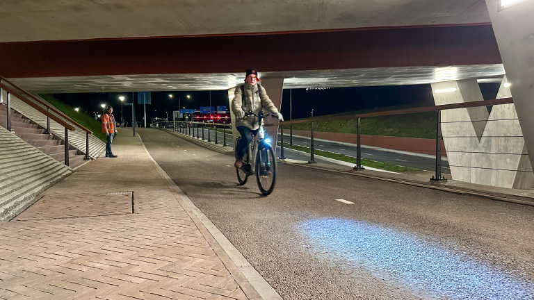 Een persoon fietst 's avonds onder een viaduct door, terwijl een andere persoon in een oranje jas toekijkt.