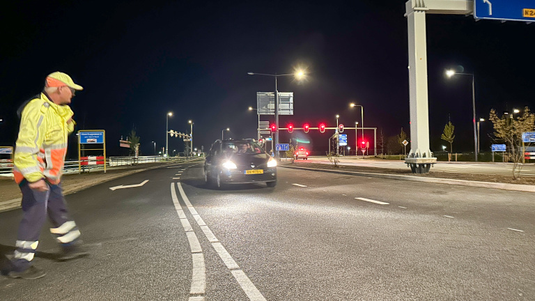 Een verkeersregelaar loopt op een weg bij nacht, terwijl een auto met brandende koplampen nadert. Verkeerslichten op de achtergrond.