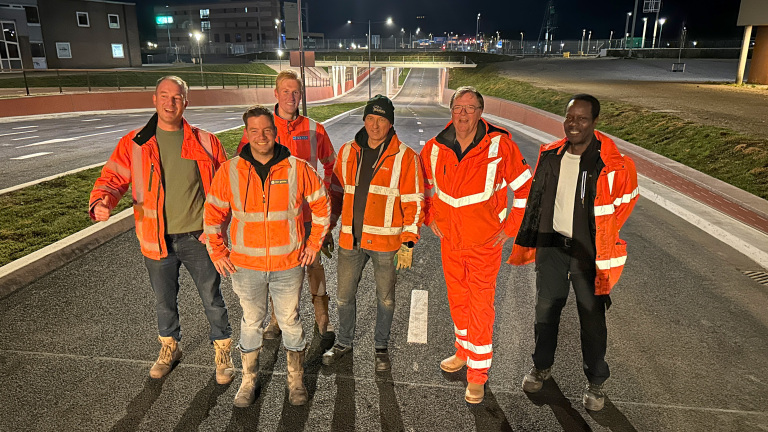 Zes mannen in feloranje veiligheidskleding staan lachend op een lege weg bij nacht, met gebouwen en lichten op de achtergrond.