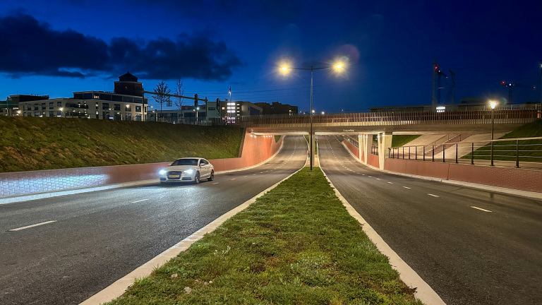 Een auto rijdt 's avonds op een verlichte weg met een viaduct en gebouwen op de achtergrond.