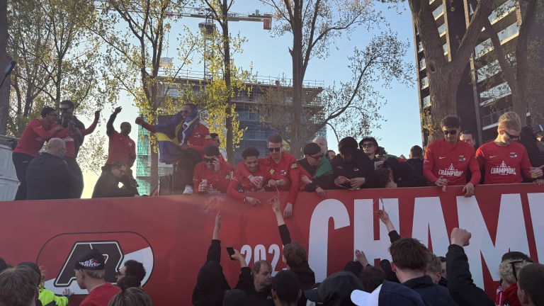 Mensen in rode shirts vieren feest op een tribune met de tekst "Champion", omringd door fans die juichen.