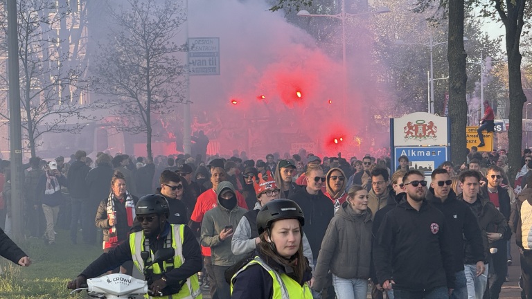 Een menigte mensen loopt door een straat in Alkmaar, omgeven door rode rook en fakkels, waaronder verkeersregelaars.
