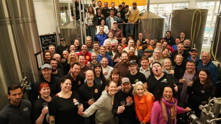 Een grote groep mensen poseert vrolijk samen in een brouwerij, velen met een drankje in de hand.