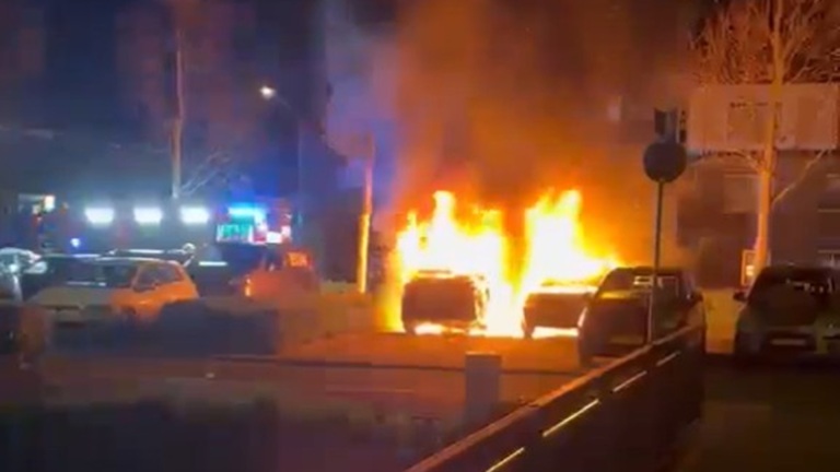 Brandende auto's in een parkeerplaats 's nachts, met vlammen en rook opstijgend naast geparkeerde voertuigen.