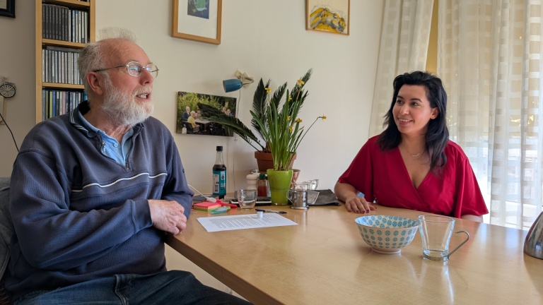 Een oudere man en een vrouw aan een tafel met kopjes, een kom en een plant, in gesprek bij het raam met schilderijen aan de muur.