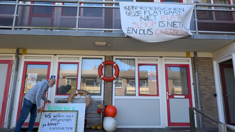 Man plaatst een bord voor een flat, met tekst tegen sloop; spandoek aan balkon met vergelijkbare boodschap.