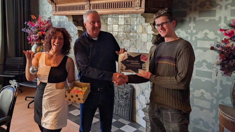 Drie mensen poseren lachend; een man schudt handen met een jongere man terwijl een vrouw een doos en een kerstbal vasthoudt.