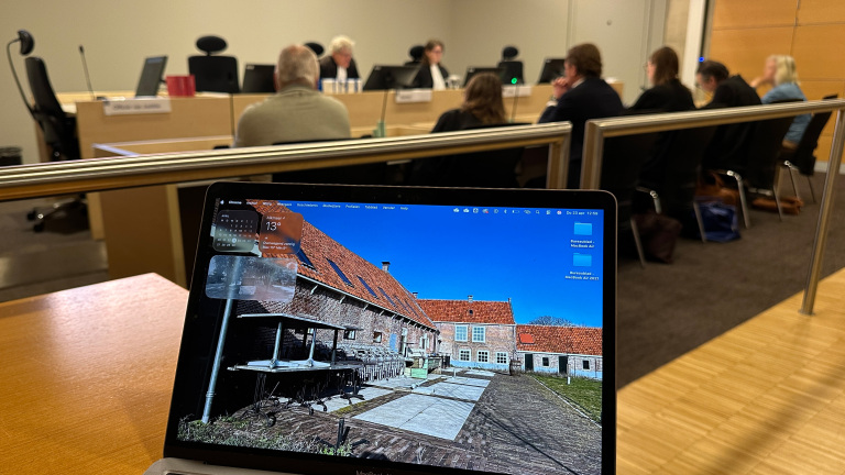 Een laptop met een foto van een gebouw op het scherm staat op een tafel in een vergaderzaal, waar mensen rond een tafel zitten.
