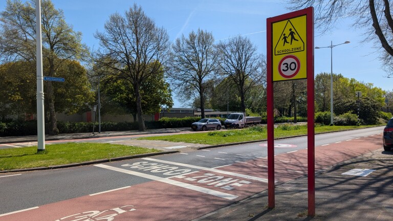 Verkeersbord met "Schoolzone" en maximumsnelheid van 30 km/u, langs een rustige straat met bomen en geparkeerde auto's.
