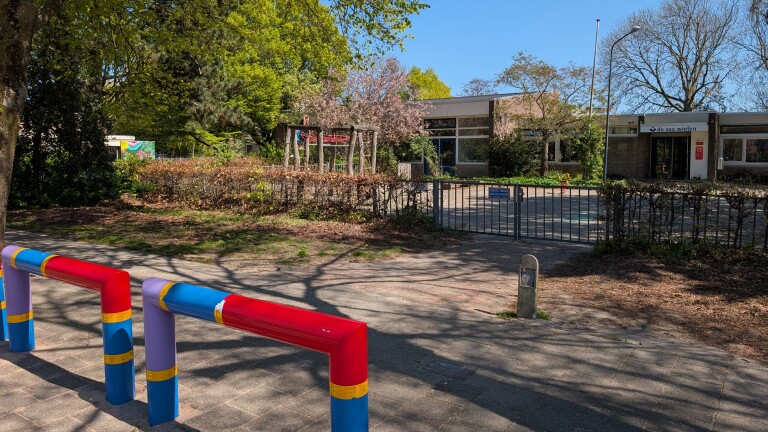 Schoolgebouw omringd door bomen, kleurrijke palen op de voorgrond en een hek voor de ingang met de tekst "de zes wielen".