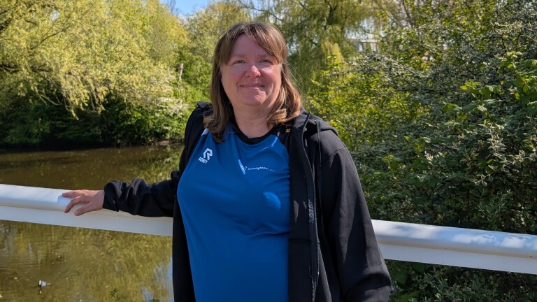Vrouw in een blauwe sportshirt leunt op een brugleuning, met groene bomen en een waterpartij op de achtergrond.