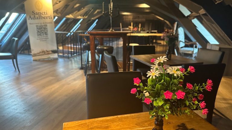 Bloemen op een tafel in een sfeervolle ruimte met houten balken, een poster van Sancti Adalberti en verschillende zitplaatsen.