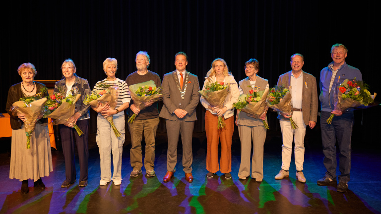 Een groep van acht mensen poseert met bloemen op een podium, naast een man in een kostuum met een ketting.