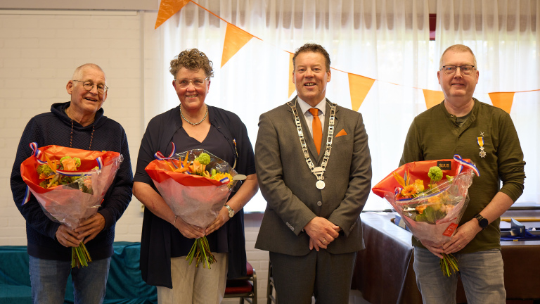Vier mensen staan naast elkaar, drie met bloemen en onderscheidingen, voor feestelijke oranje versiering.