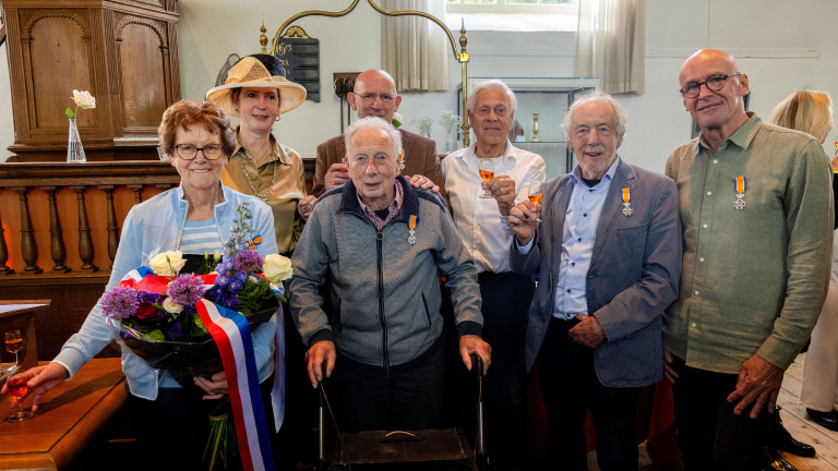 Groep mensen in een zaal, sommigen met onderscheidingen en bloemen, houden glazen in de lucht voor een viering.