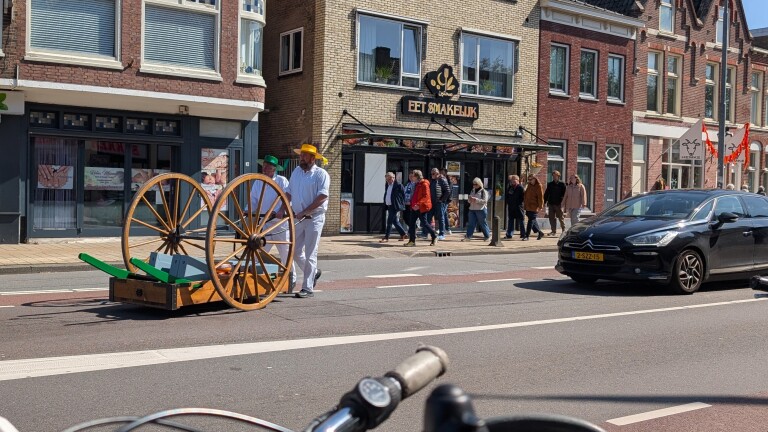 Twee mannen in witte kleding met gekleurde hoeden duwen een houten kaaskar voor een winkel, omringd door voetgangers en auto's.