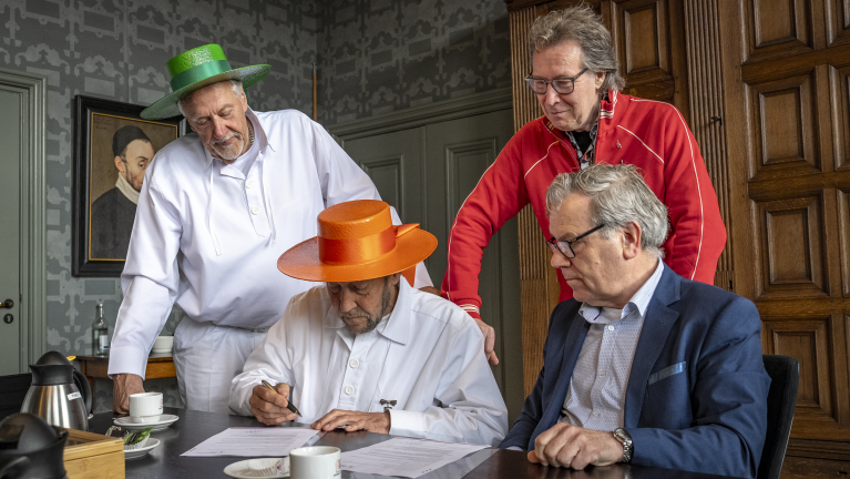 Vier mannen in een kamer, waarvan er twee kleurrijke hoeden dragen, zitten aan tafel terwijl een van hen een document ondertekent.