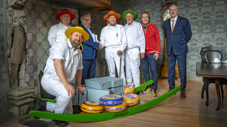 Groep mannen in kleurrijke hoeden en witte kleding poseren met kazen op een drager in een historische kamer.