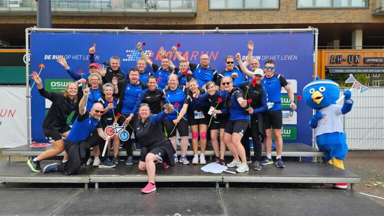 Een groep mensen in sportkleding poseert enthousiast met rode bloemen, naast een blauw mascotte kostuum.