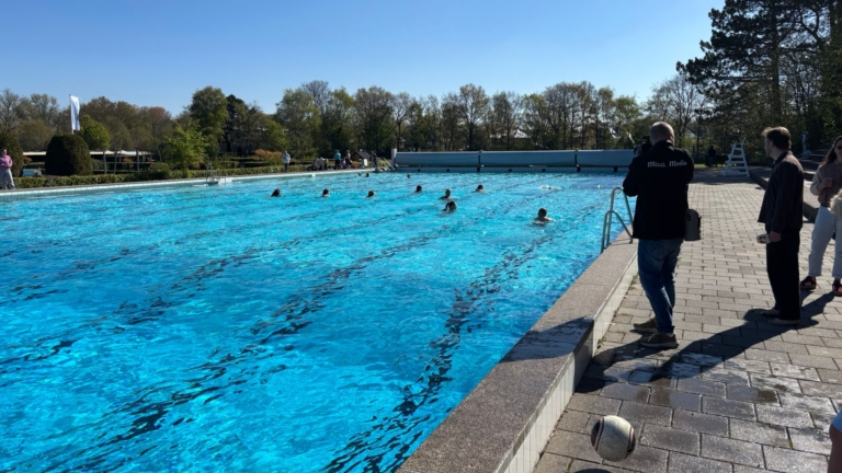 Zwembad met mensen die zwemmen op een zonnige dag, omringd door bomen en toeschouwers langs de rand.