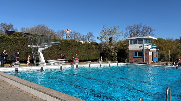 Een jongen springt van een duikplank in een buitenzwembad op een zonnige dag, omringd door mensen die toekijken.