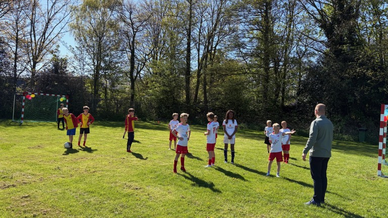 Kinderen spelen voetbal op een grasveld, begeleid door een volwassene bij zonnig weer.