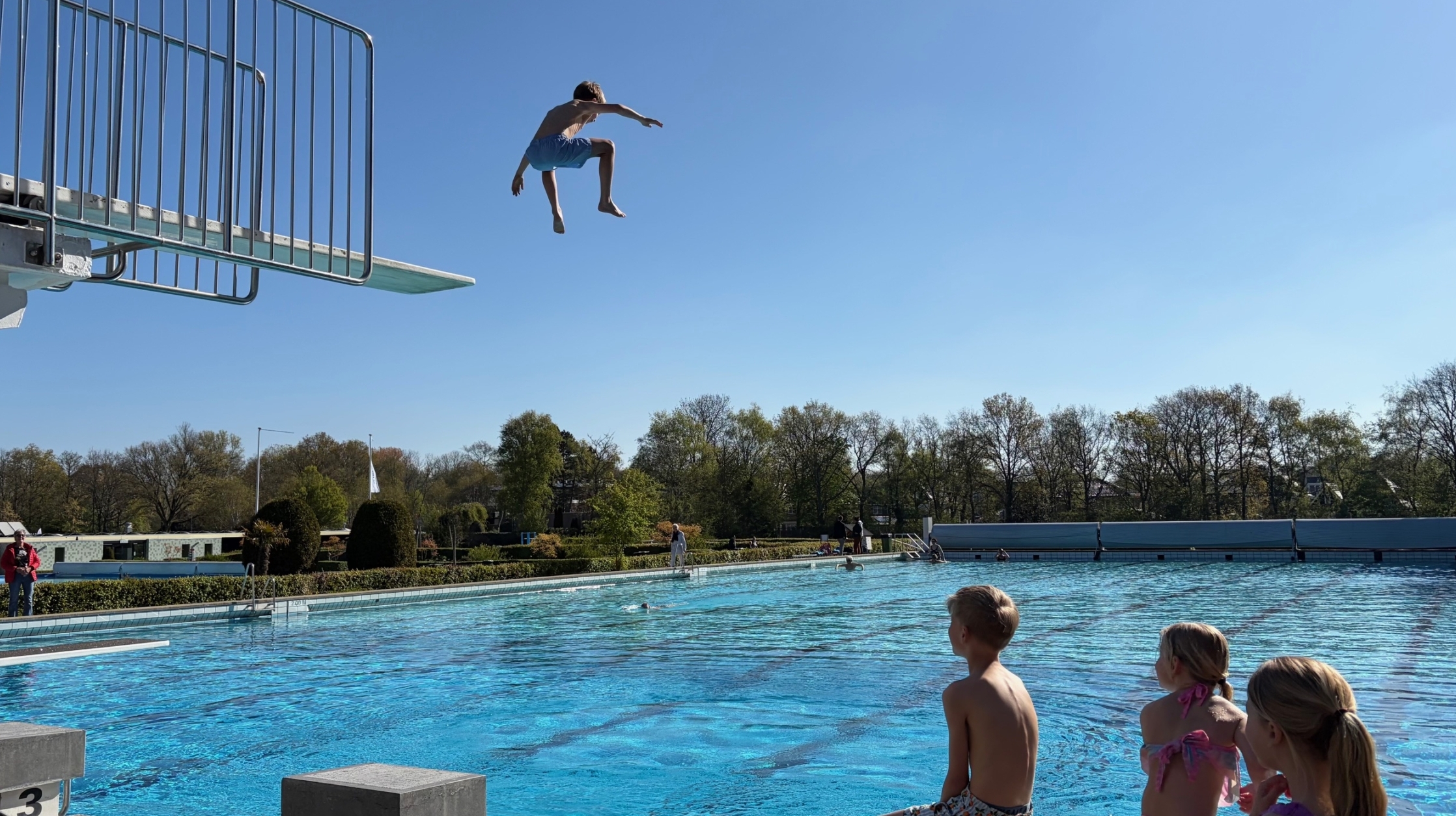 Een jongen springt van een hoge duikplank in een buitenzwembad, terwijl drie kinderen toekijken.