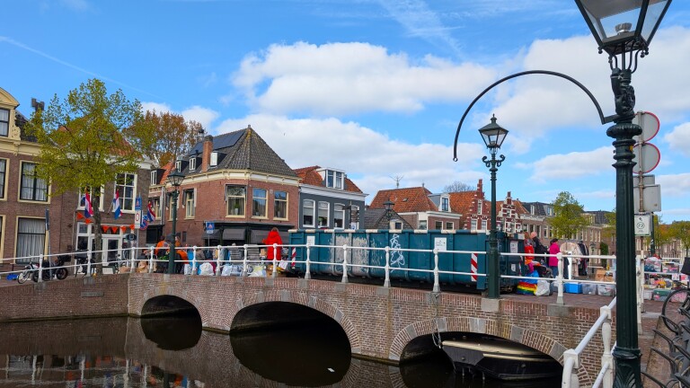 Een stenen brug met mensen langs kraampjes en versieringen, omgeven door traditionele Nederlandse huizen onder een blauwe lucht.