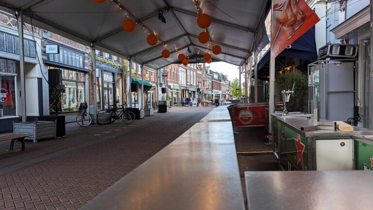 Straatbeeld met winkels aan beide zijden, een overdekte zithoek met oranje decoratie en een biertap met Amstel-logo.