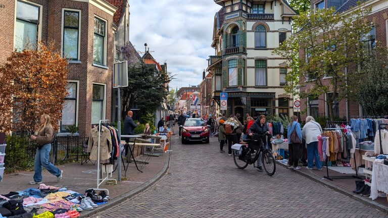 Straat met mensen die langs kraampjes met kleding en andere items lopen tijdens een buurtmarkt, met huizen op de achtergrond.
