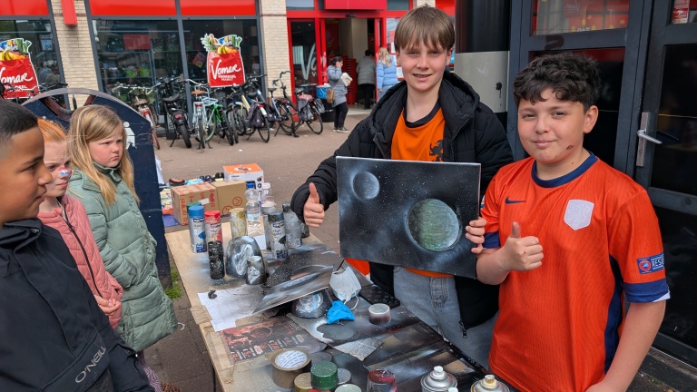 Kinderen bij een marktkraam met spuitbuskunst; twee jongens tonen trots een schilderij van planeten.