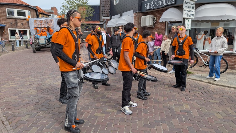 Jonge drummers in oranje shirts spelen op straat tijdens een parade, met een versierde tractor op de achtergrond.