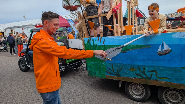 Man in oranje jas neemt deel aan een feestelijke parade terwijl een jongen hem iets reicht met een peddel.