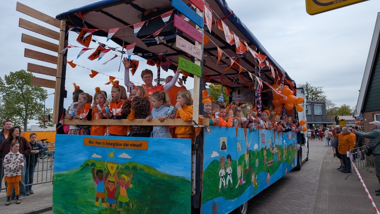 Een versierde praalwagen met kinderen gekleed in oranje, omgeven door oranje versieringen, rijdt door een straat tijdens een feestelijke optocht.