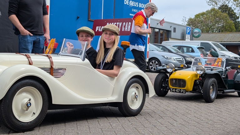 Kinderen in vintage speelgoedauto's lachen, terwijl een man op de achtergrond loopt langs geparkeerde auto's.
