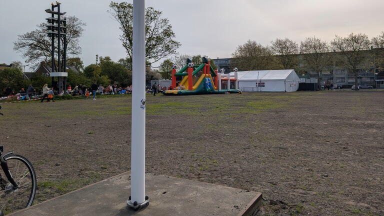 Open terrein met een springkussen en een groep mensen bij een tent, op de achtergrond bomen en woningen.