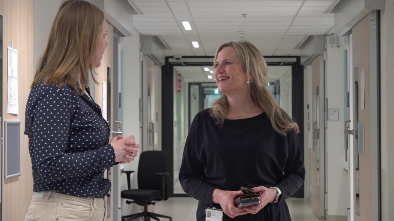 Twee vrouwen staan pratend in een goed verlichte gang van een gebouw, één met een polkadot blouse.