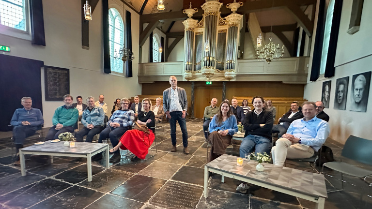 Groep mensen zittend in een kerkachtige ruimte met een groot orgel op de achtergrond, ontspannen en glimlachend.