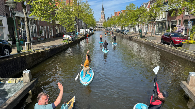 Mensen peddelen op SUP-boards langs een historische gracht met bomen en stenen gebouwen aan weerszijden, op een zonnige dag.