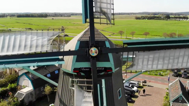 Windmolen met radars en wieken in een groen landschap, met huizen en geparkeerde auto's op de achtergrond.