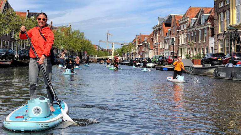 Mensen suppen door een gracht met emmers op hun boards, omgeven door historische gebouwen en een blauwe lucht.