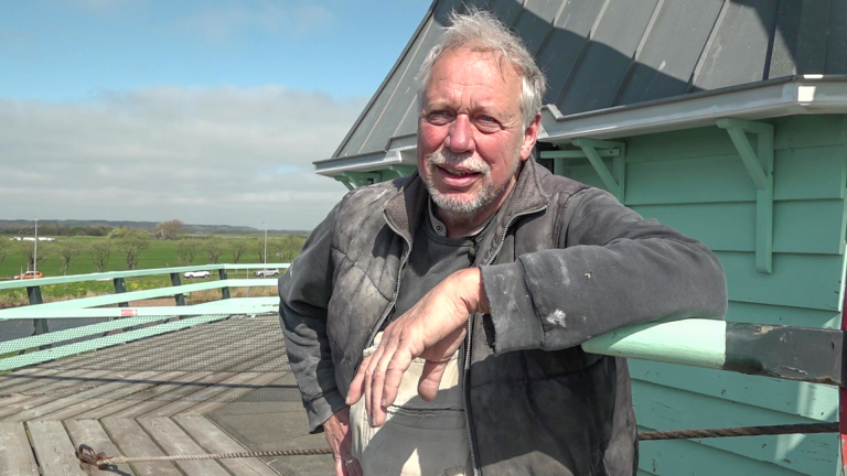 Man met grijs haar en baard leunt op een railing bij een gebouw met groene houten gevel, met een weids landschapszicht.