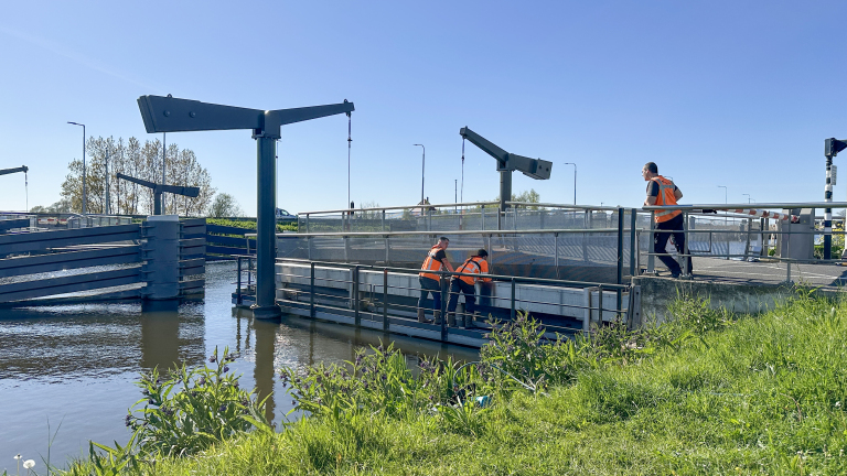 Twee arbeiders in oranje hesjes werken bij een sluis met een heldere blauwe lucht op de achtergrond.