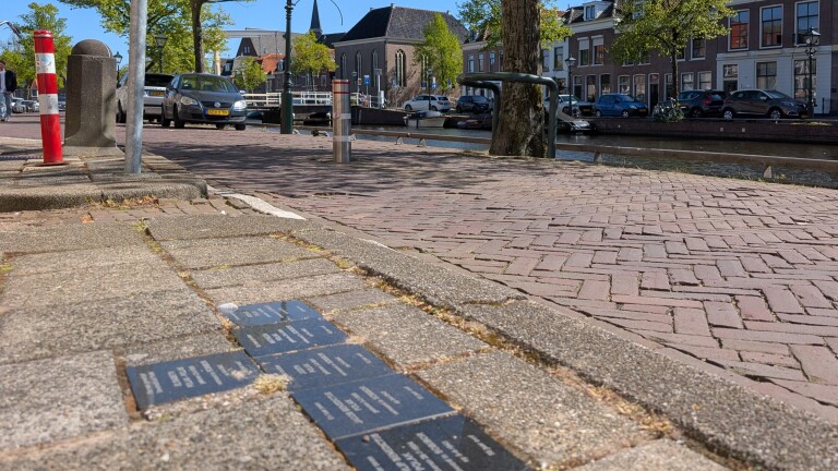 Gedenkstenen ingebed in een trottoir naast een straat en kanaal, met bomen en geparkeerde auto's in de achtergrond.