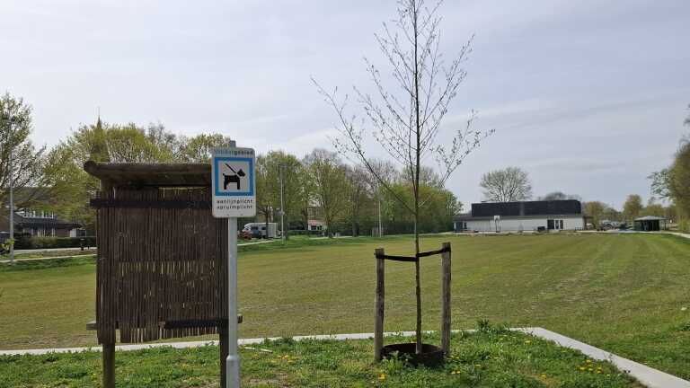 Bord met tekst "Uitlaatgebied" en pictogram van een hond aan de lijn, op een grasveld met enkele bomen en gebouwen op de achtergrond.