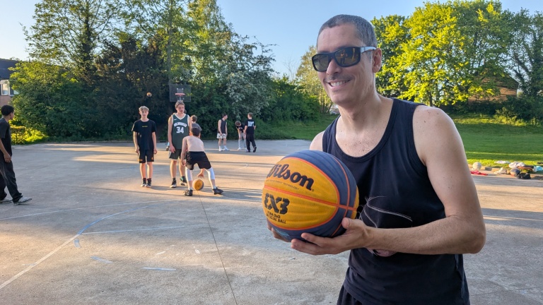 Man met zonnebril houdt een basketbal vast, terwijl op de achtergrond mensen basketballen op een buitenveld.