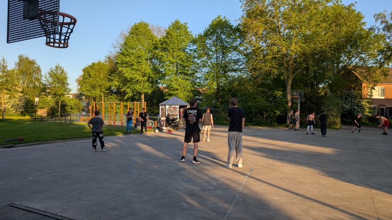 Mensen spelen basketbal op een buitenterrein omgeven door bomen op een zonnige dag.